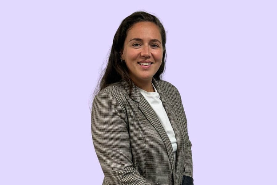 Photo de Mélanie Da Silva, jeune femme brune sur font mauve aux couleurs d'Hoppen. Veste professionnelle, sourire confiant, elle respire la confiance acquise dans son expertise de chef de projet portail patient chez Hoppen