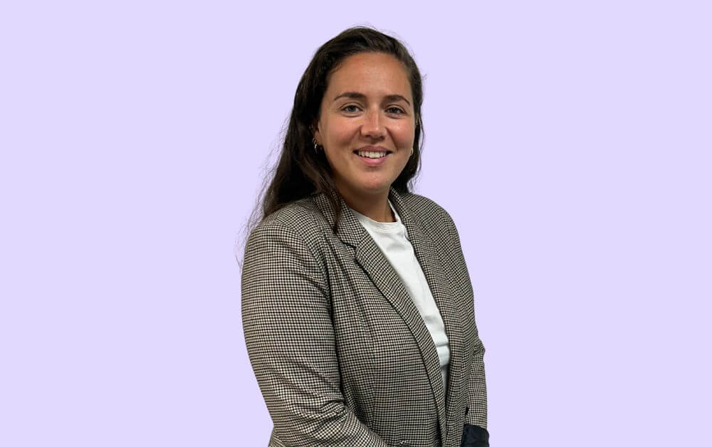 Photo de Mélanie Da Silva, jeune femme brune sur font mauve aux couleurs d'Hoppen. Veste professionnelle, sourire confiant, elle respire la confiance acquise dans son expertise de chef de projet portail patient chez Hoppen