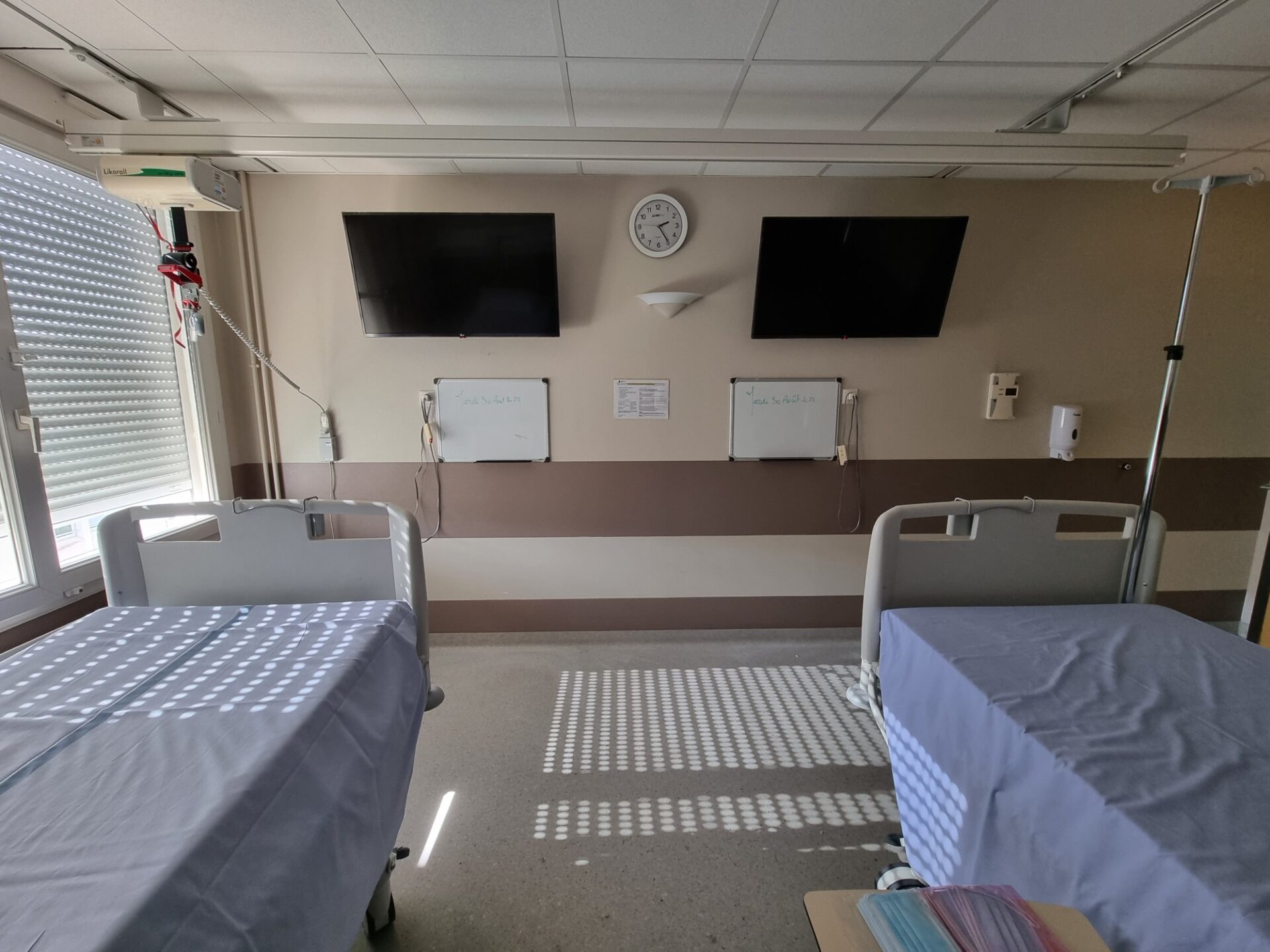 Photo de fin de déploiement en établissement. On voit 2 lits d'hôpital en chambre double, avec les 2 tableaux de communication médicale et 2 écrans installés par Hoppen dans la lumière qui filtre à travers des stores.